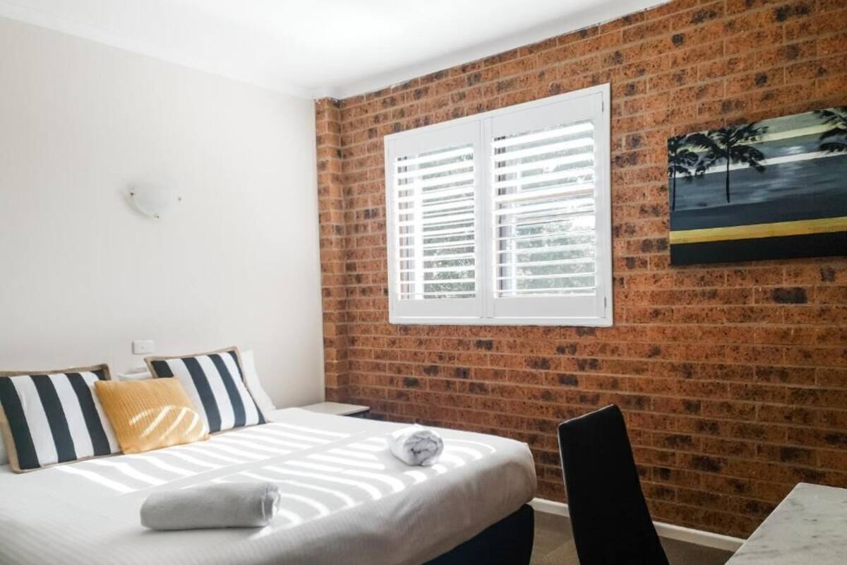 Habitación de hotel con pared de ladrillo, cama con almohadas a rayas, ventana con contraventanas y un cuadro de una puesta de sol.