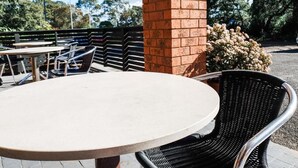 Un espacio al aire libre con una mesa redonda y sillas de mimbre, un pilar de ladrillo y un fondo de árboles y una cerca.