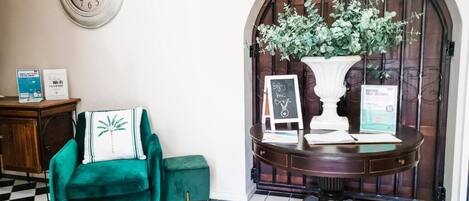 A checkered floor, a green armchair, a round table with a vase of flowers, a clock, and a door with a sign.