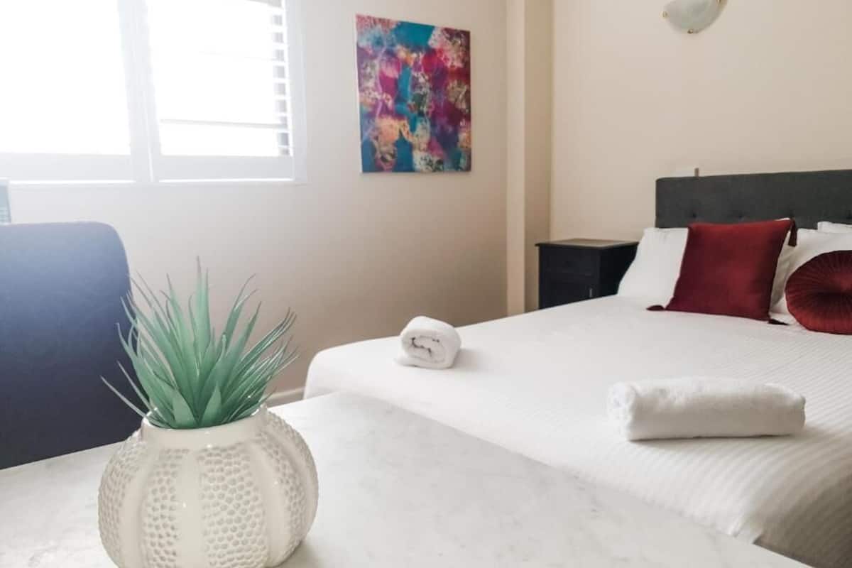 Una habitación de hotel con una cama, una mesita de noche con una planta, una toalla enrollada, un cuadro abstracto colorido y una lámpara.