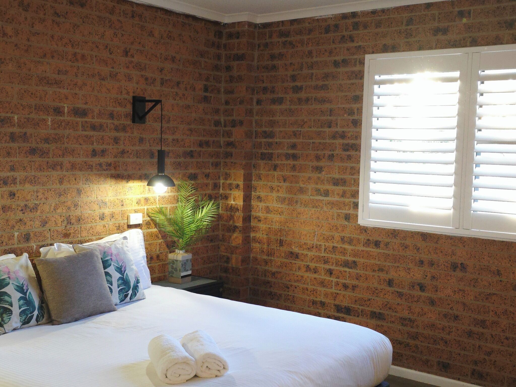 Une chambre d’hôtel avec un mur en briques, un lit recouvert de draps blancs, une plante en pot, une table de chevet avec une lampe et une fenêtre avec des stores.