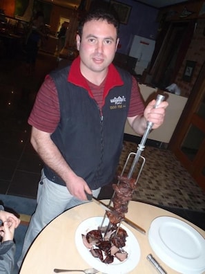 A person standing at a table with a meat skewer, a plate with meat slices, and a fork. - Snow Gate Motel and Apartments (Berridale)