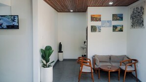 A modern living room with a wooden ceiling, a beige sofa, two wooden chairs, a small round table, and a flat-screen TV mounted on the wall. - Batu Kandik Homestay (Pecatu)