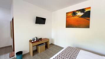 Quarto de hotel com cama, escrivaninha, cadeira, lixeira, televisão e um quadro na parede.
