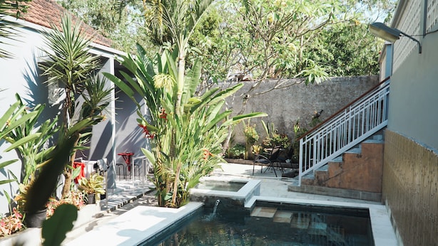 A swimming pool with a small waterfall feature, surrounded by potted plants and a staircase leading to a balcony. - Batu Kandik Homestay (Pecatu)