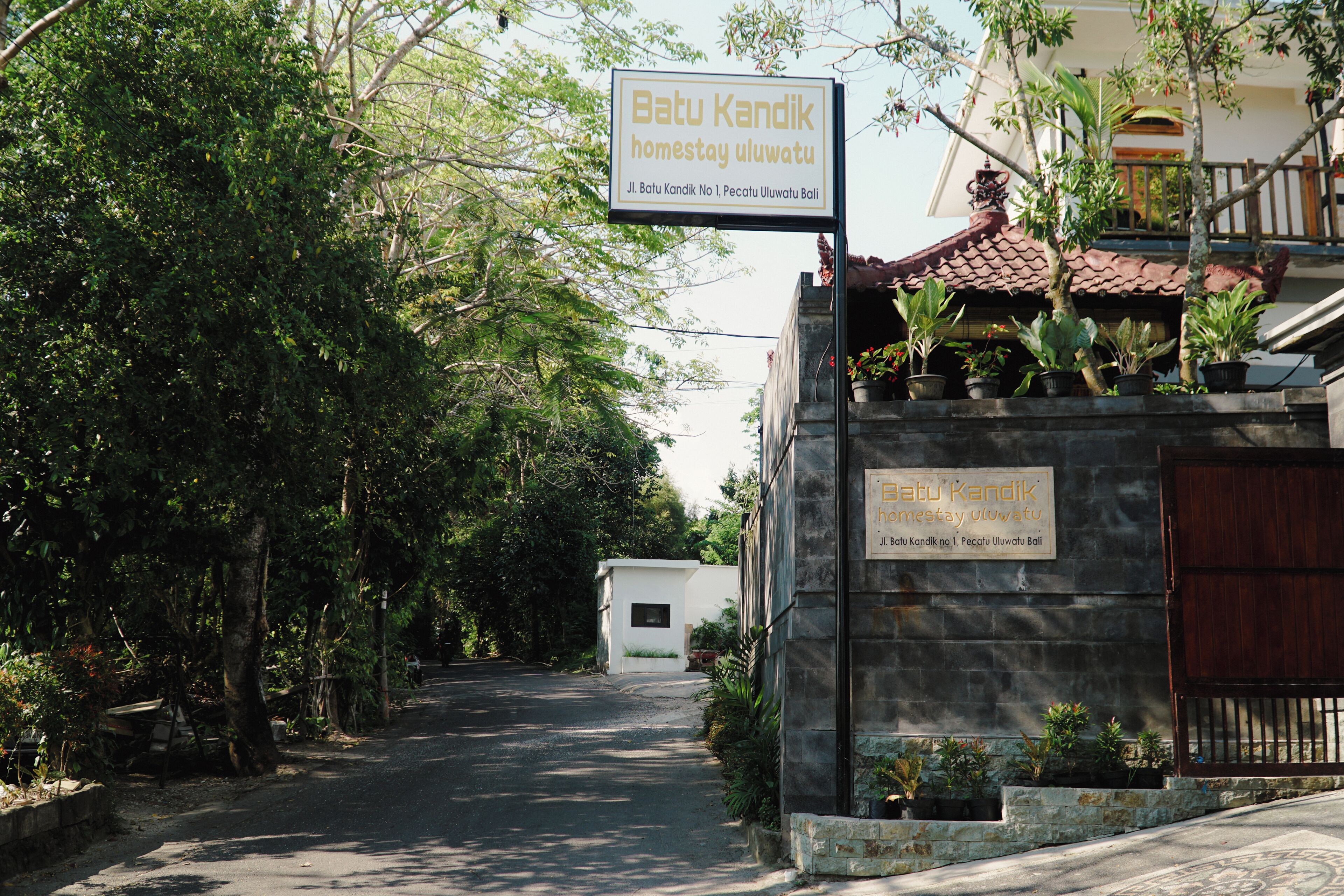 A sign for 'Batu Kandik homestay uluwatu' with a view of a road leading to a building with a tiled roof.