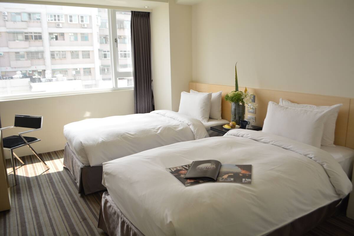 A hotel room with two beds, a chair, a nightstand, and a window with a view of buildings.