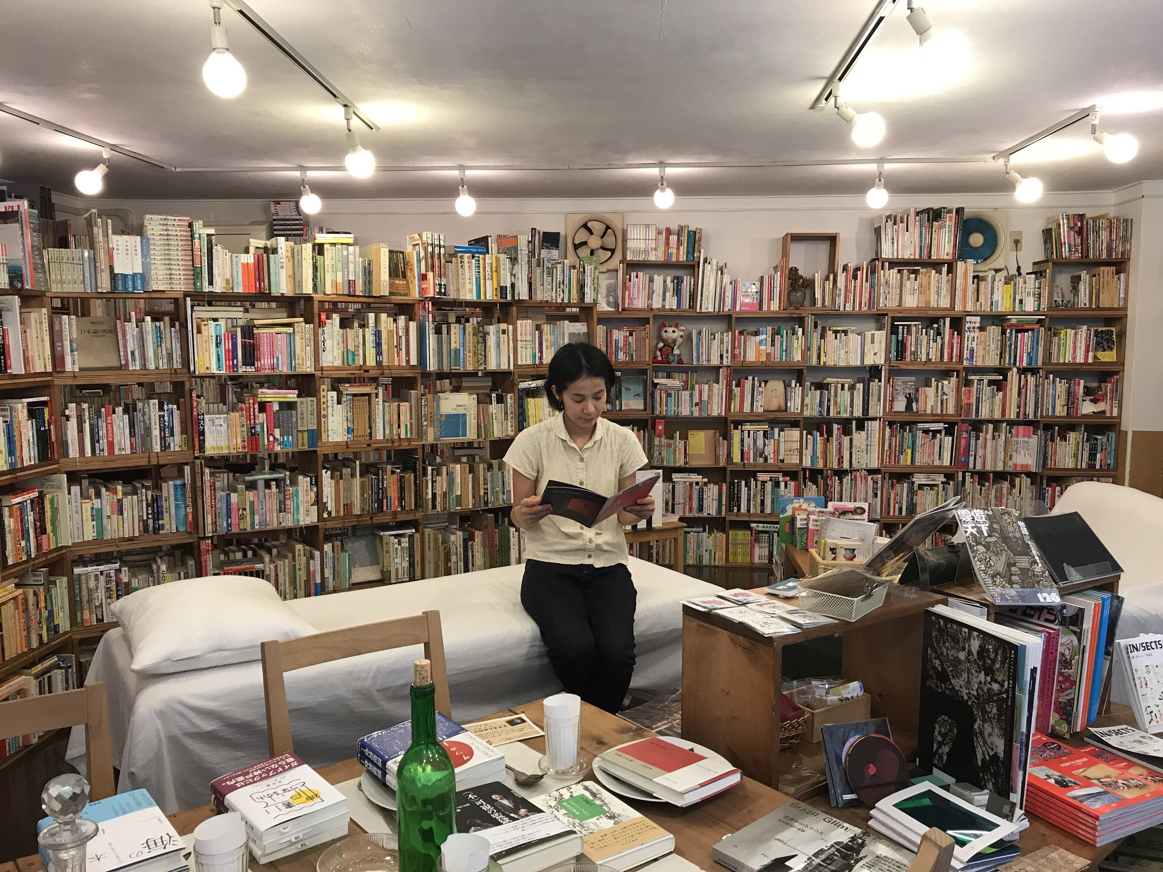 Une bibliothèque avec des étagères remplies de livres, un coin lecture avec un lit, et une table sur laquelle se trouvent des livres et une bouteille.