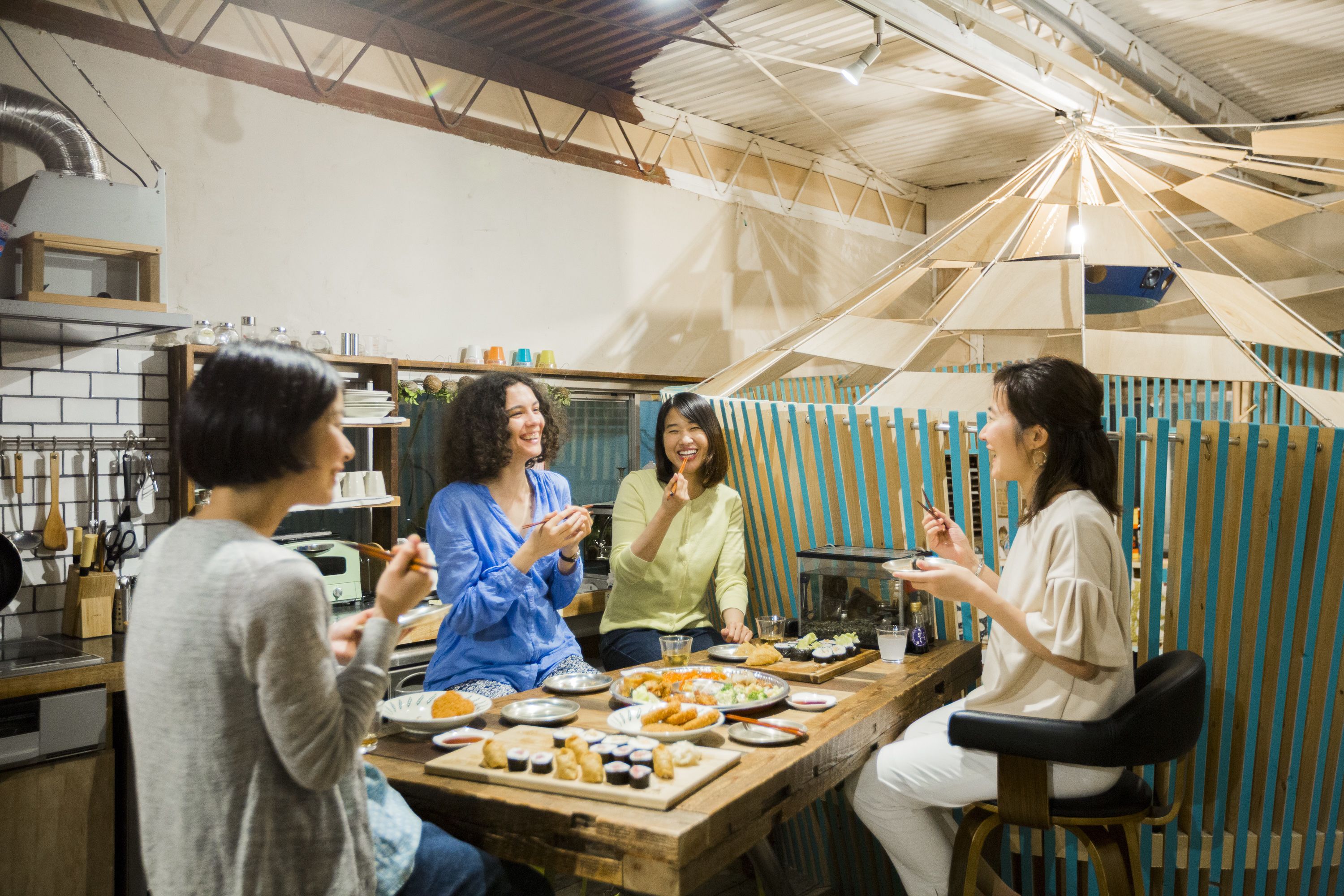 A group of people dining at a wooden table in a rustic kitchen setting with a high ceiling and exposed beams.