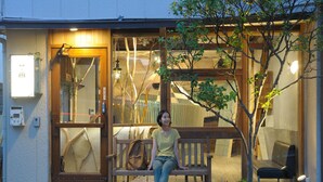 A person sitting on a wooden bench outside a building with a glass door and a small tree in front. - Guest House MAYA - Hostel (Kobe)