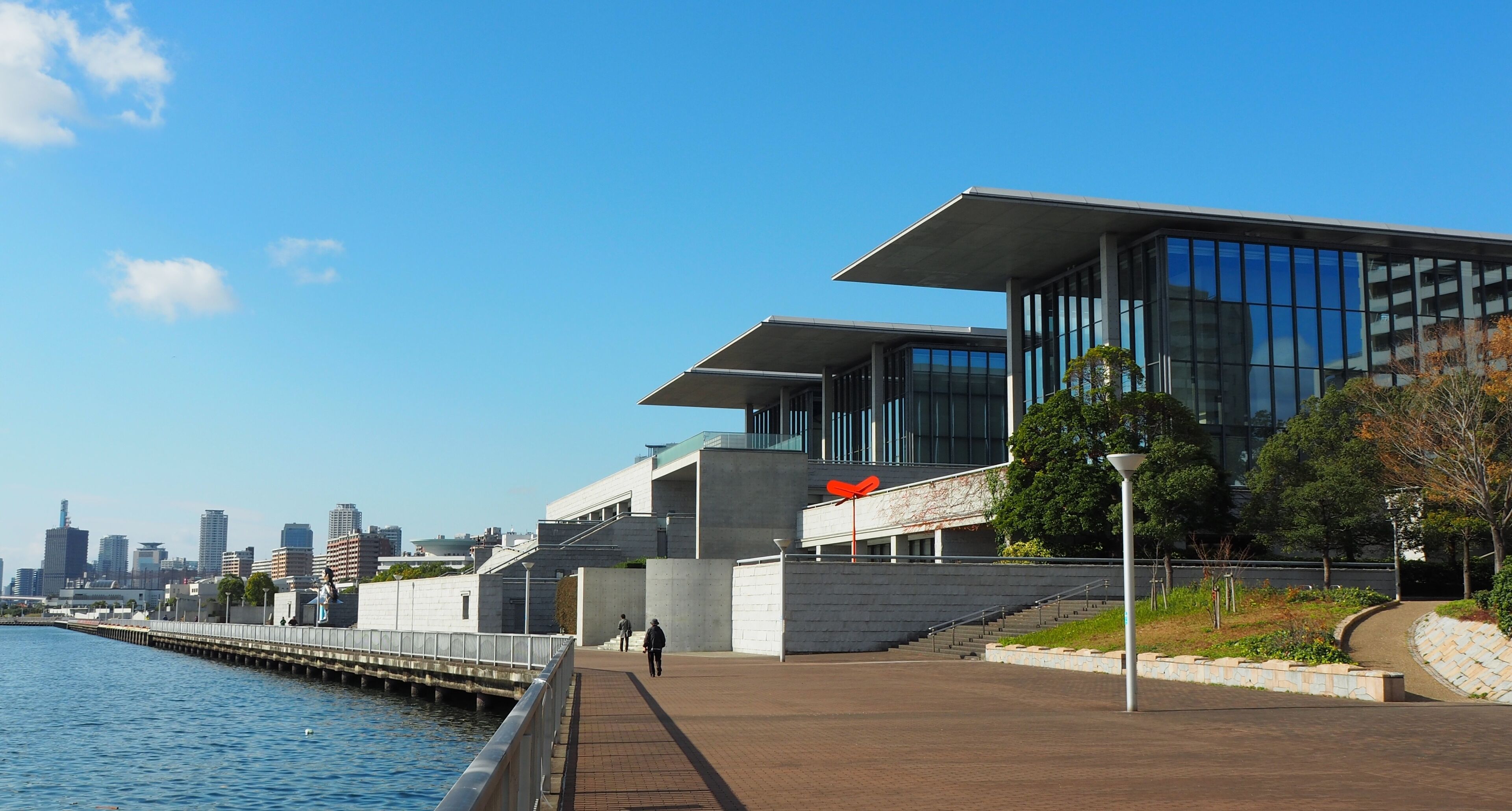 A waterfront walkway with modern buildings, a clear sky, and a city skyline in the background.