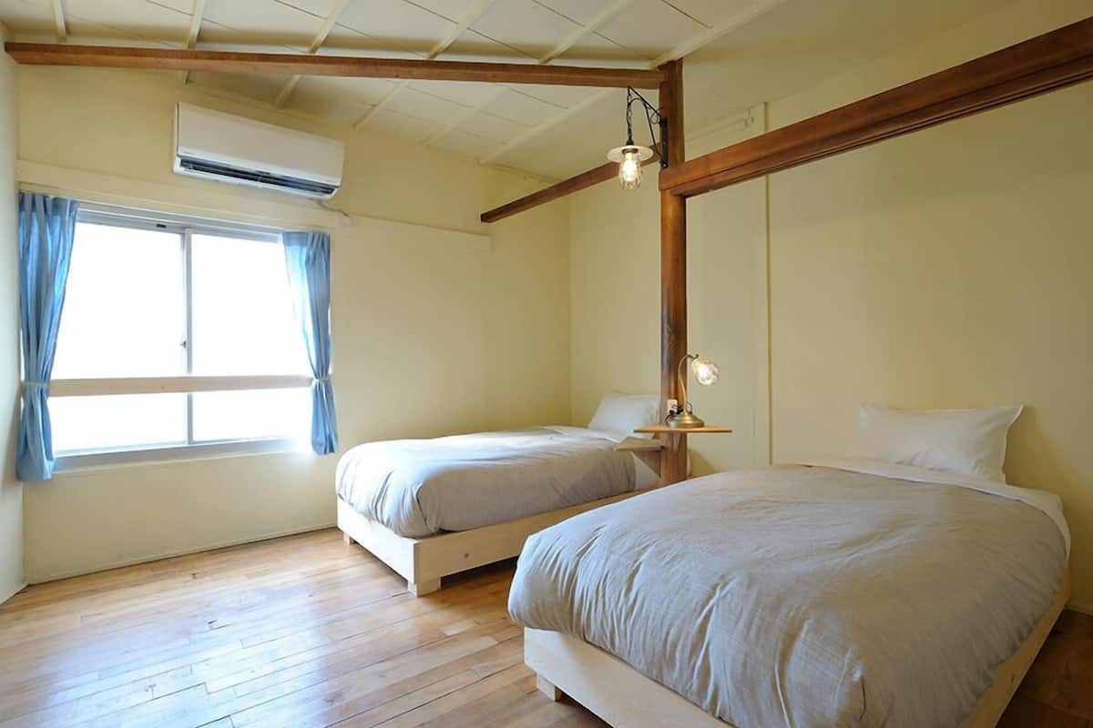 A bedroom with two beds, wooden flooring, a window with curtains, an air conditioner, and a hanging light fixture.