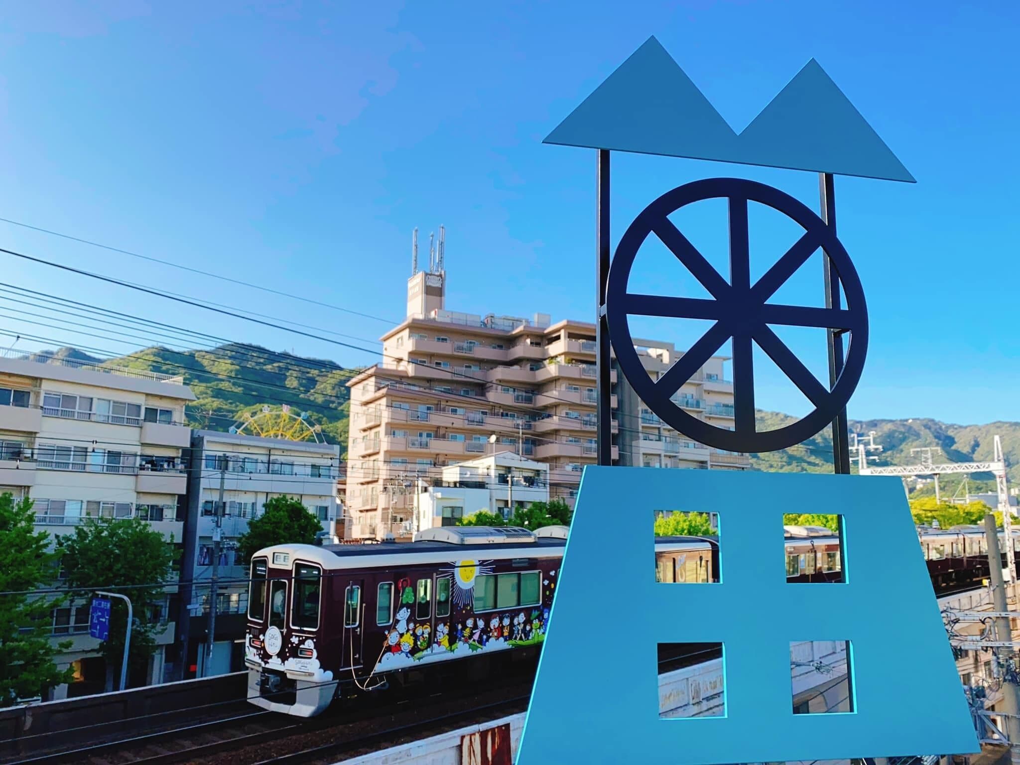 A train with a colorful exterior, passing by a building with a large wheel symbol and a blue structure with windows.