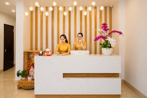 A reception desk with two staff members, a wooden slat wall, hanging lights, and a potted orchid.