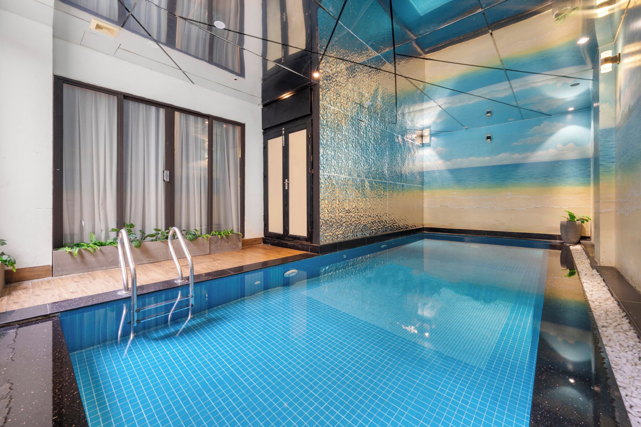 Una piscina interior con baldosas azules cristalinas, una pared de vidrio con un mural de playa y una escalera metálica para entrar.