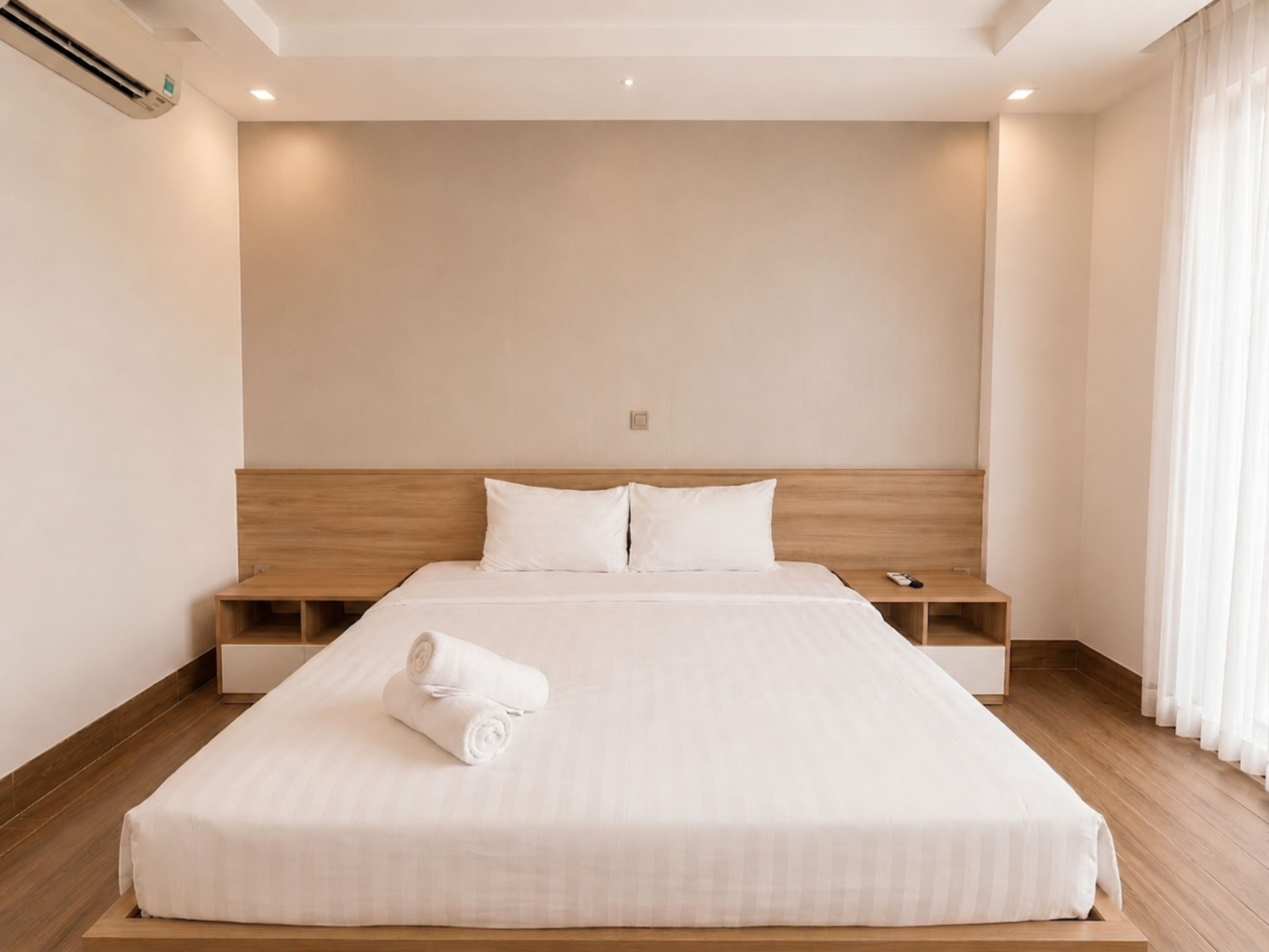 Habitación de hotel con una cama grande, cabecera de madera, dos mesitas de noche y una toalla enrollada sobre la cama.