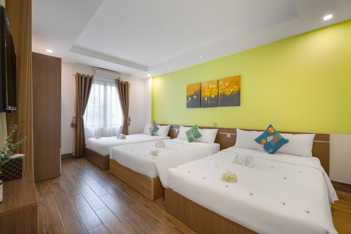 Ein Hotelzimmer mit drei Betten, einem großen Fenster mit Vorhängen, einer grünen Akzentwand und Kunstwerken über den Betten.