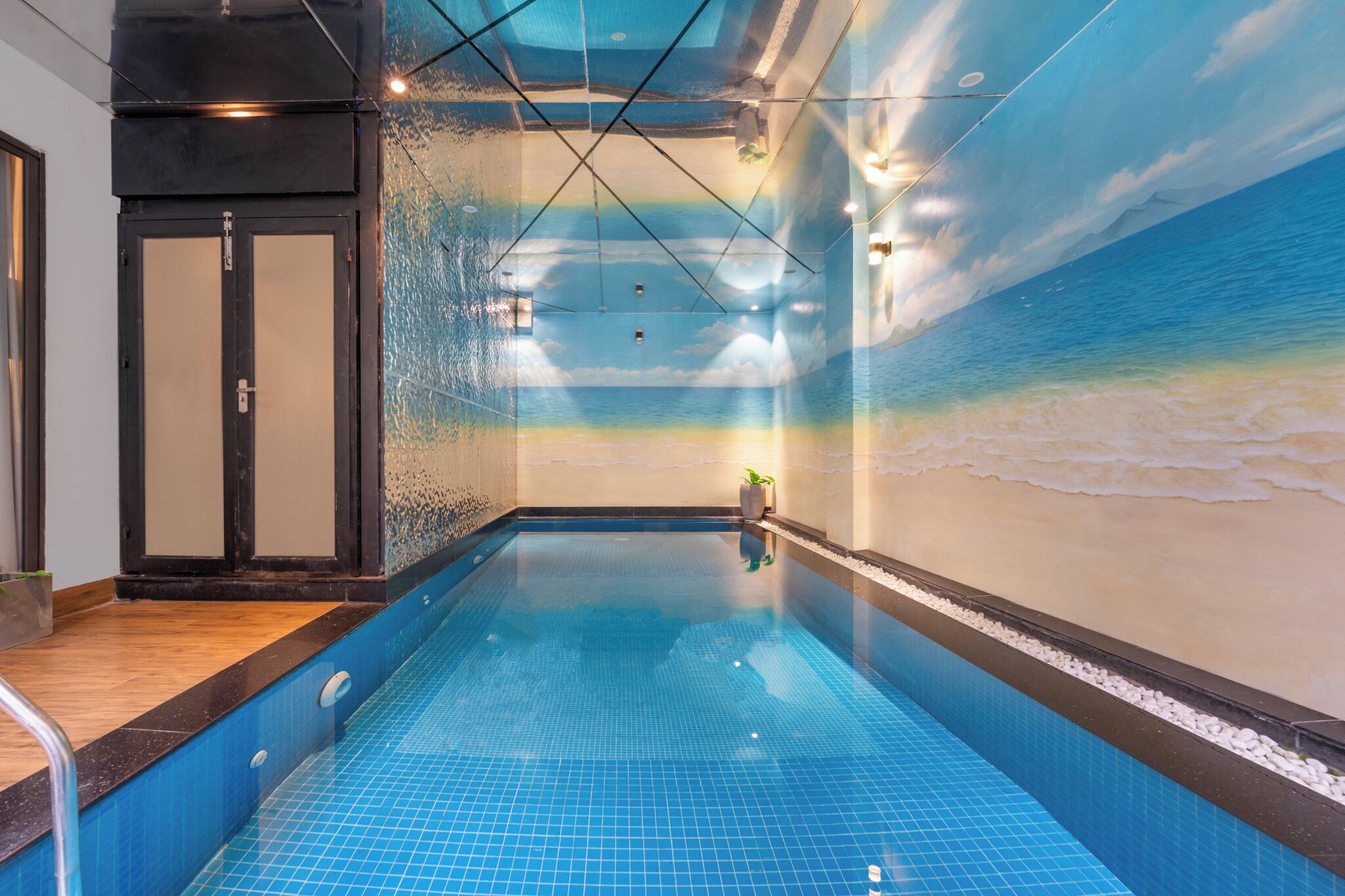 Una piscina interior con un mural de playa en la pared, una zona de terraza de madera y una persona parada junto a la piscina.