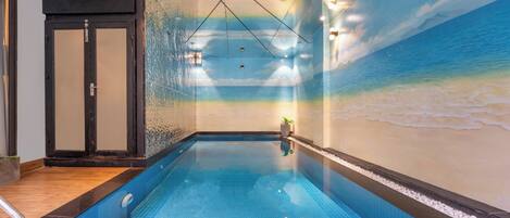 Una piscina interior con un mural de playa en la pared, una zona de terraza de madera y una persona parada junto a la piscina.