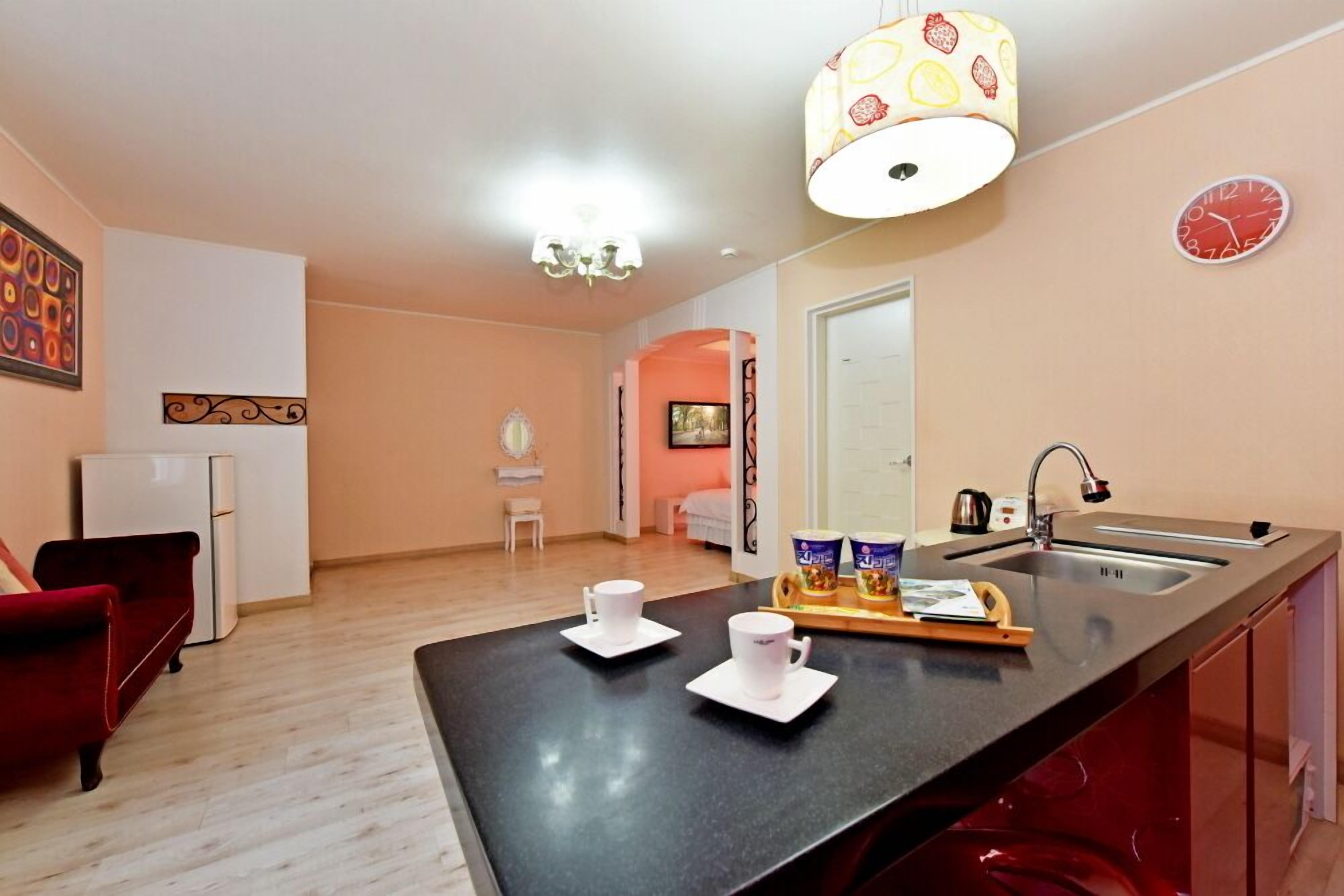 Una cocina moderna con encimera negra, un refrigerador, un lavabo y un comedor con una silla roja y dos tazas blancas.