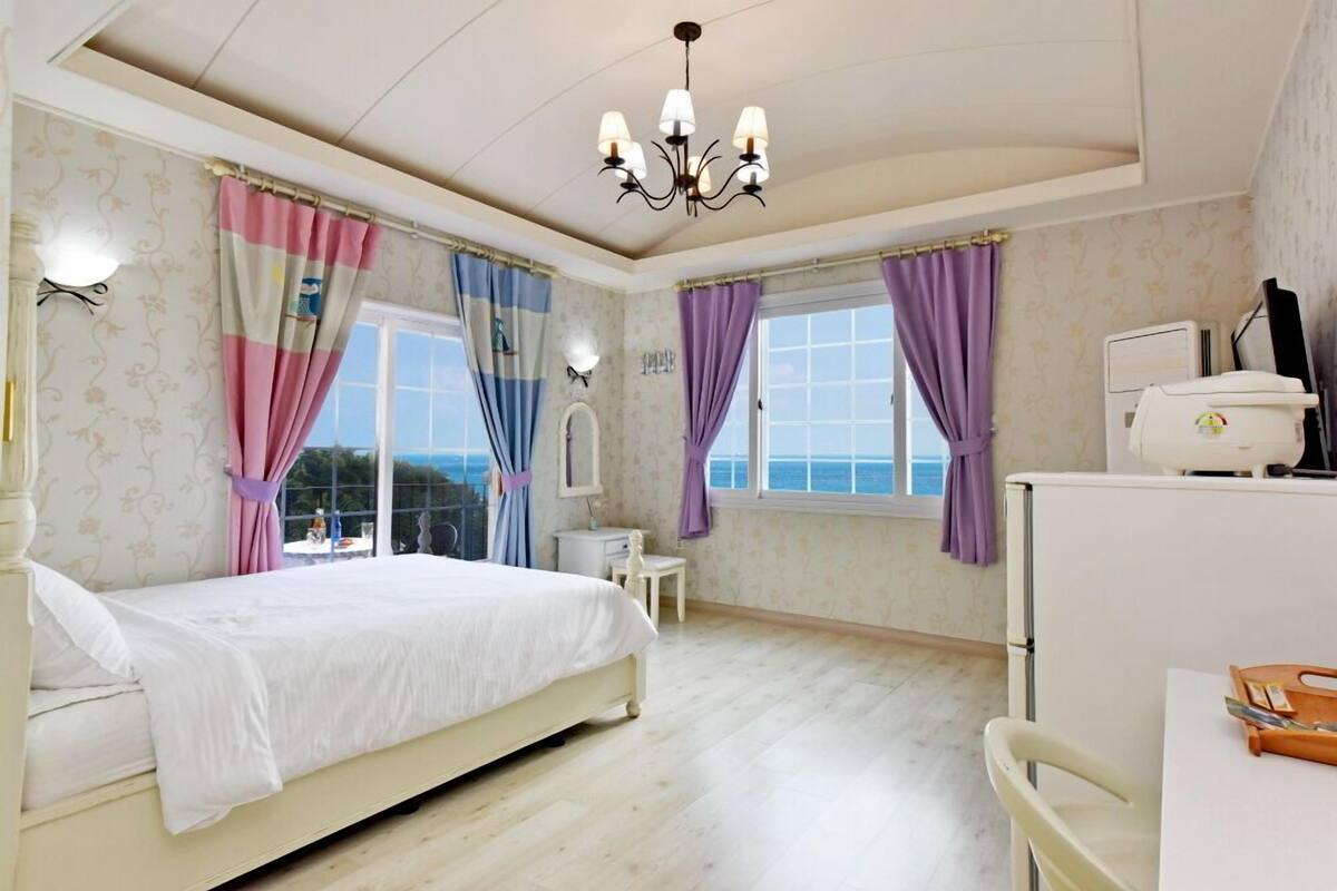Ein Schlafzimmer mit Bett, Schreibtisch, Stuhl, einem Fenster mit Vorhängen, einer Chandelieber und Blick aufs Meer.