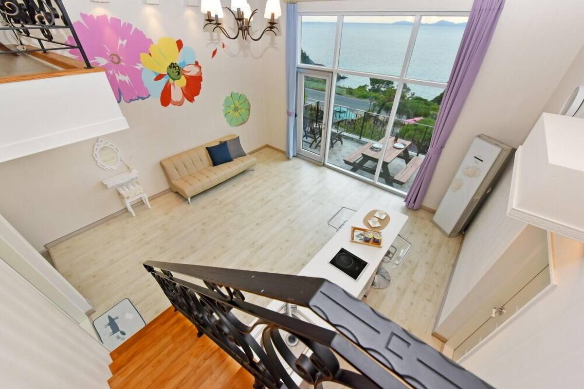 Ein geräumiger Wohnbereich mit großem Fenster mit Blick auf einen Pool und das Meer, eine Couch, ein Esstisch und eine Treppe, die zu einer oberen Etage führt.