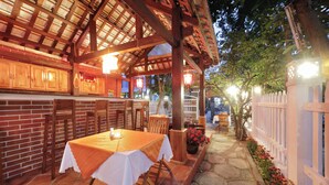 An outdoor patio with wooden tables and chairs, a brick wall, and a white fence with flowers. - Ly Phuc Homestay (Da Nang)