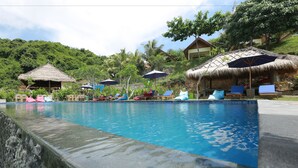 A pool area with lounge chairs and umbrellas, surrounded by thatched-roof structures and lush greenery. - Artati Lombok Bungalows & Restaurant (Selong Belanak)