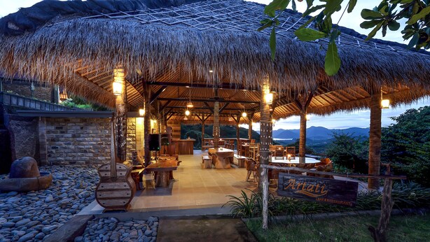 An outdoor dining area with a thatched roof, stone walls, and a view of the mountains. - Artati Lombok Bungalows & Restaurant (Selong Belanak)