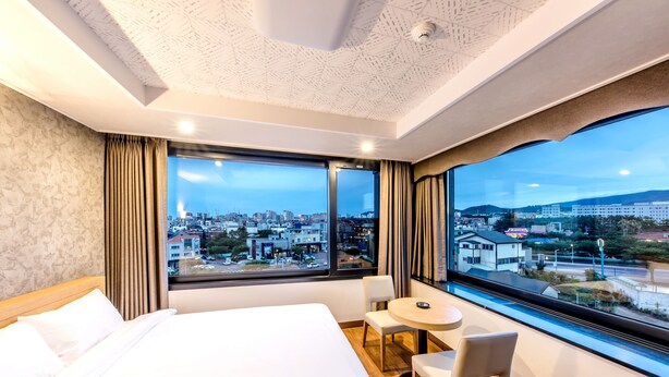 Une chambre d’hôtel avec un grand lit, une table ronde et une vue sur le paysage urbain, visible à travers de grandes fenêtres.