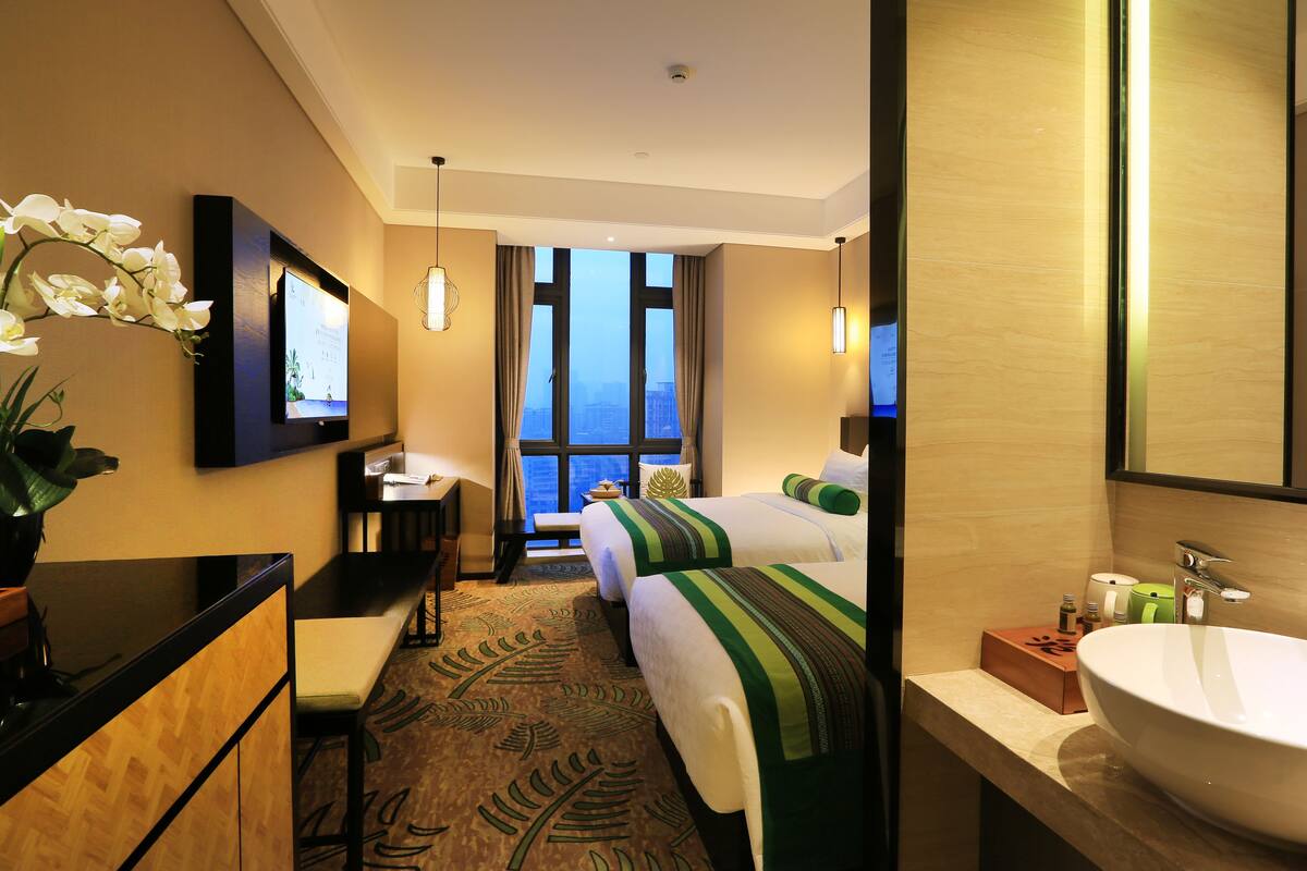 A hotel room with a bed, a desk, a chair, a large window with a city view, a bathroom with a sink and mirror, and a potted plant.
