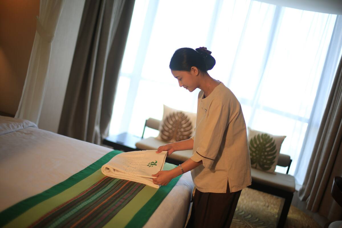 A hotel room with a neatly made bed, a green and white striped blanket, and a large window with curtains.