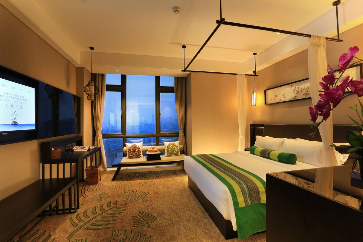 A hotel room with a large bed, a TV on the wall, a desk with a computer, and a window with a city view.