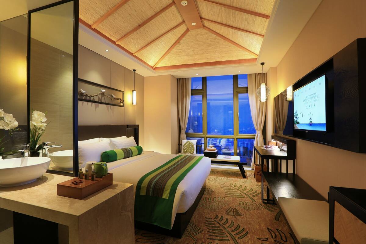 A hotel room with a large bed, a desk, a TV, a bathroom with a sink and mirror, and a view of the city through large windows.