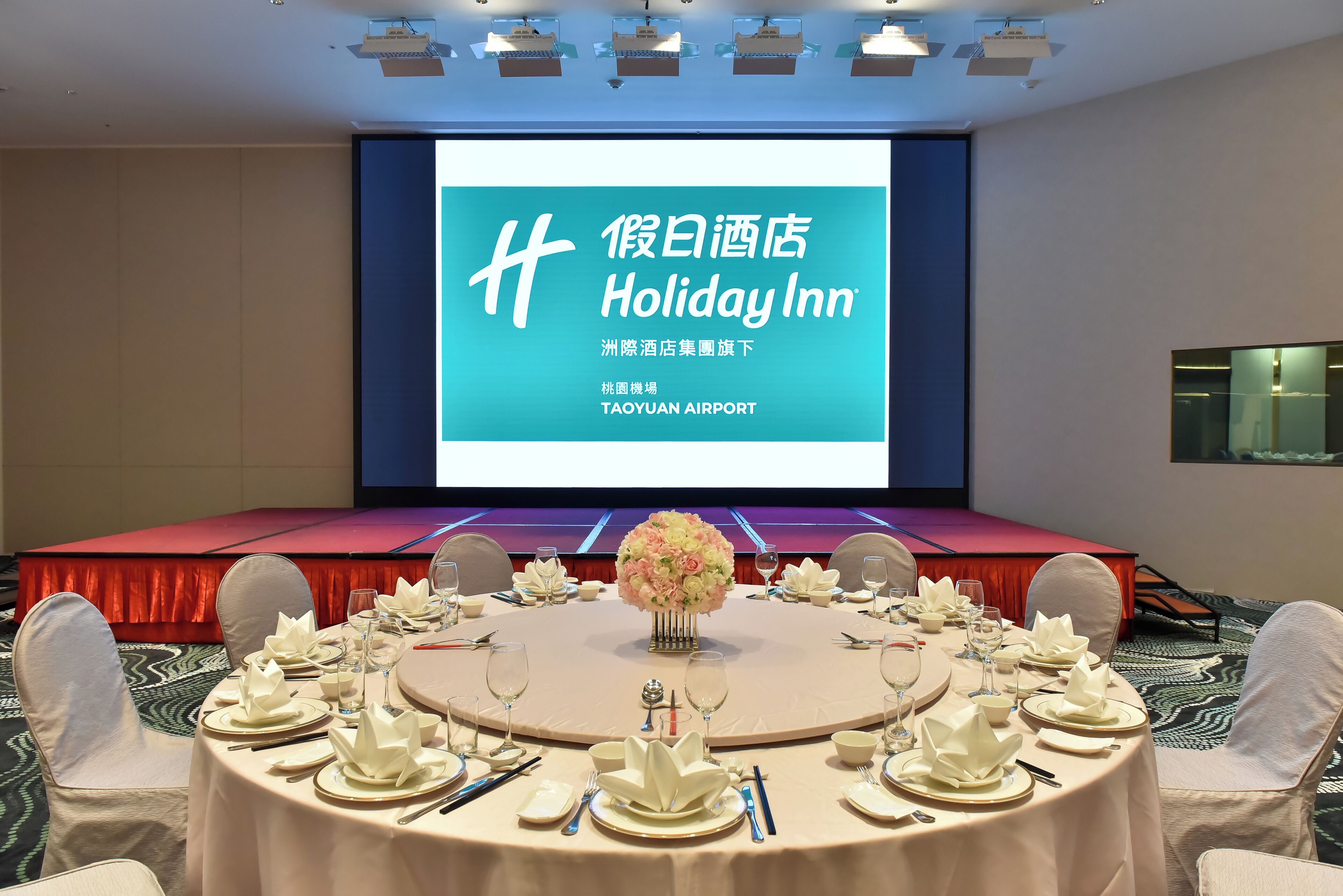 A conference room with a large screen displaying "Holiday Inn" and "TAOYUAN AIRPORT" text, surrounded by round tables set for a meeting.