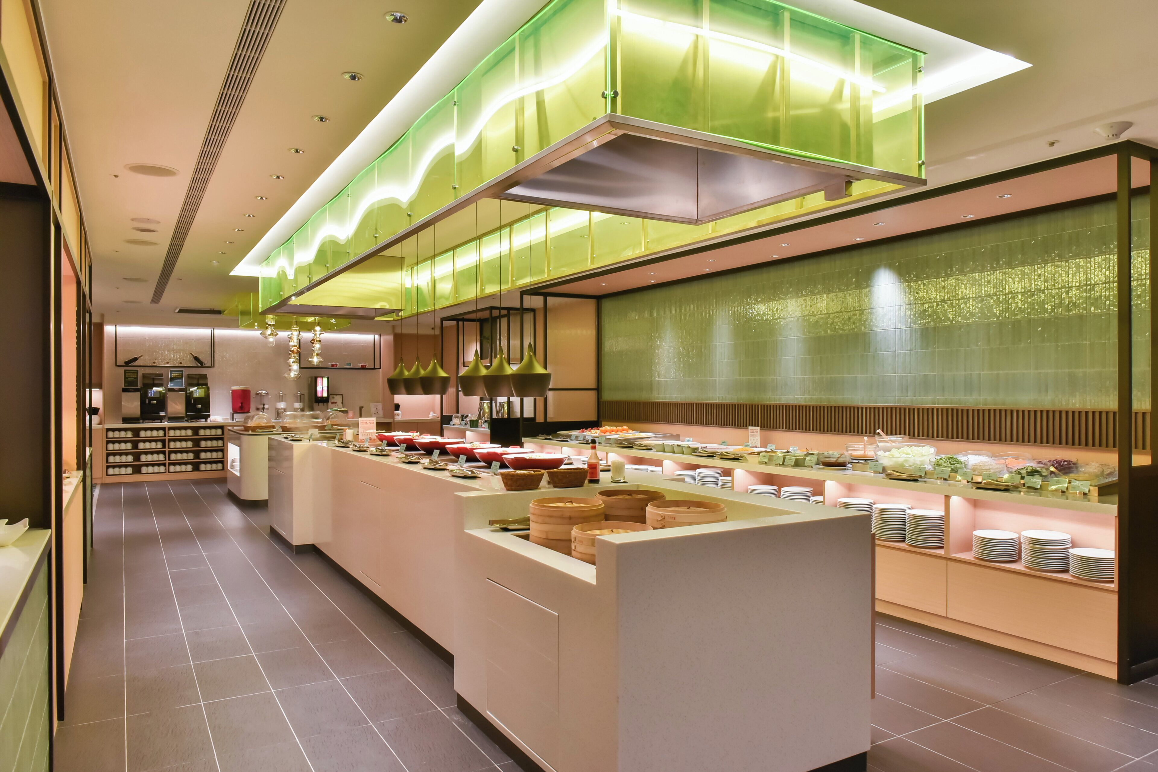 A modern hotel lobby with a central reception desk, a buffet area with various food items, and a large green illuminated panel on the ceiling.