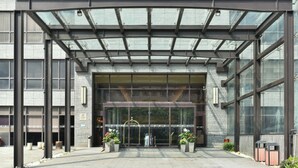 Una entrada moderna de edificio con toldo de vidrio, ventanas amplias y plantas en macetas.