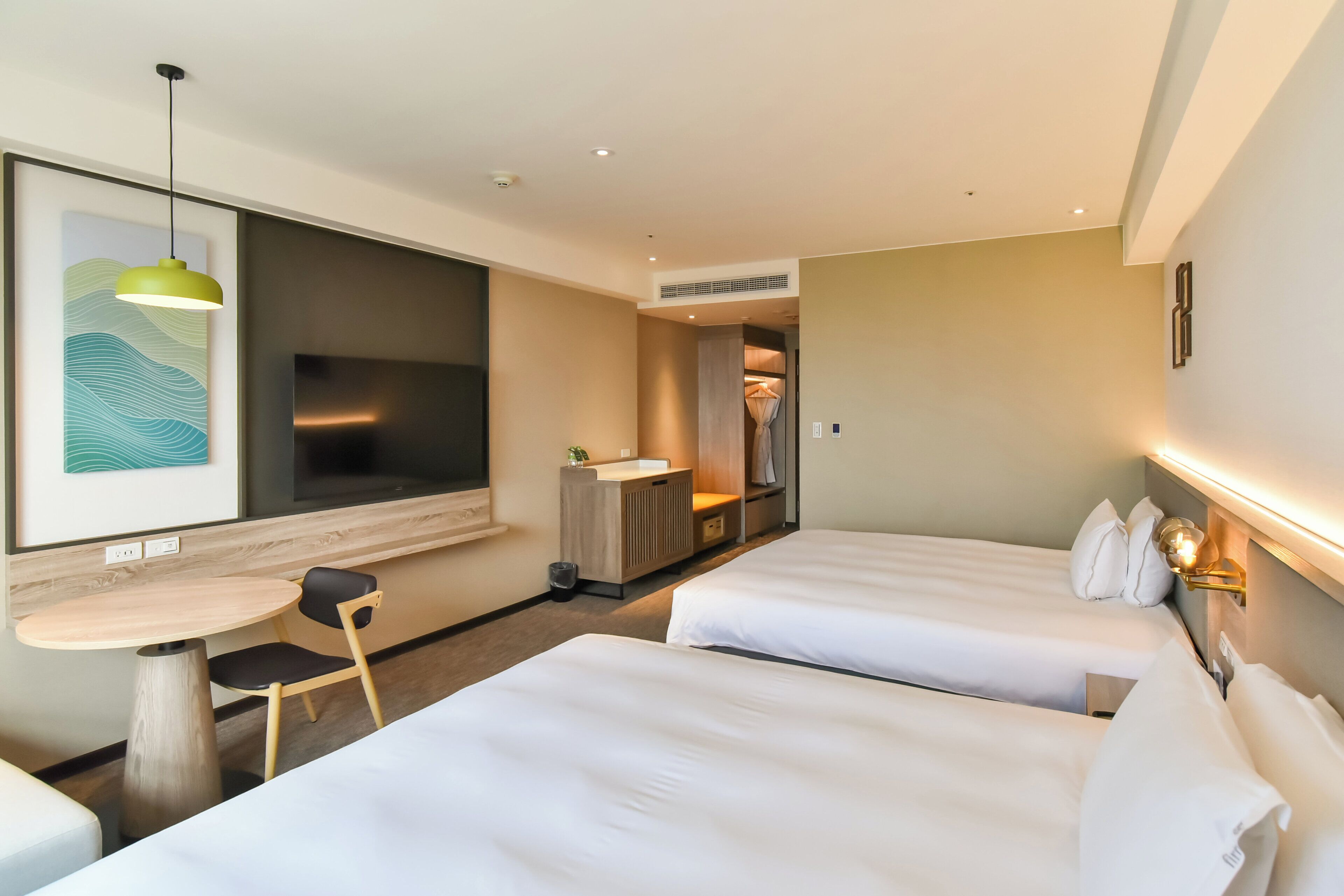 Quarto de hotel com duas camas, uma mesa pequena com uma cadeira, uma televisão e um banheiro visível por uma porta aberta.