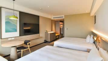 Quarto de hotel com duas camas, uma mesa pequena com uma cadeira, uma televisão e um banheiro visível por uma porta aberta.