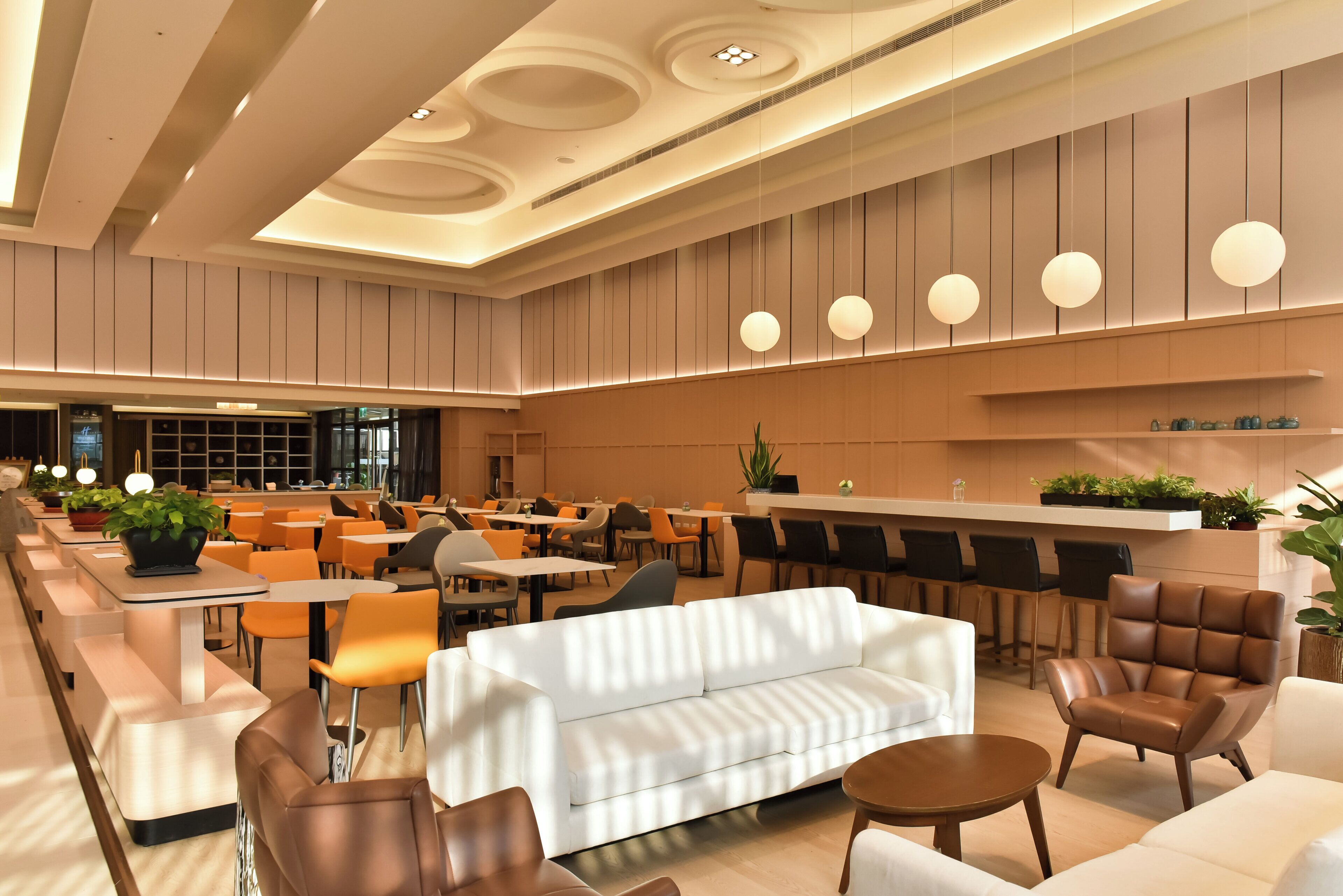 Eine geräumige Lobby mit modernem Design, ausgestattet mit einer zentralen Sitzecke, einem weißen Sofa und mehreren Sesseln, einer Bar mit Hockern sowie verschiedenen Pflanzen und dekorativen Elementen.
