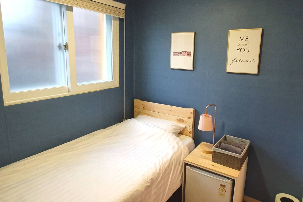 A small bedroom with a single bed, a nightstand, a lamp, a trash bin, and two framed pictures on the wall.