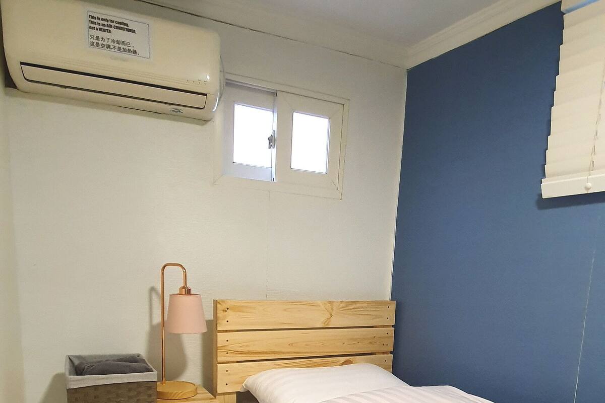 A small, single-bed room with a wooden headboard, a bedside table with a lamp, a small refrigerator, and a window with blinds.