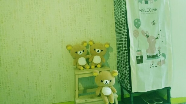 A room with a green wall, a small wooden stool, and three teddy bears. There is a welcome sign with a bear illustration on the wall. - FILL HOUSE (Seoul)