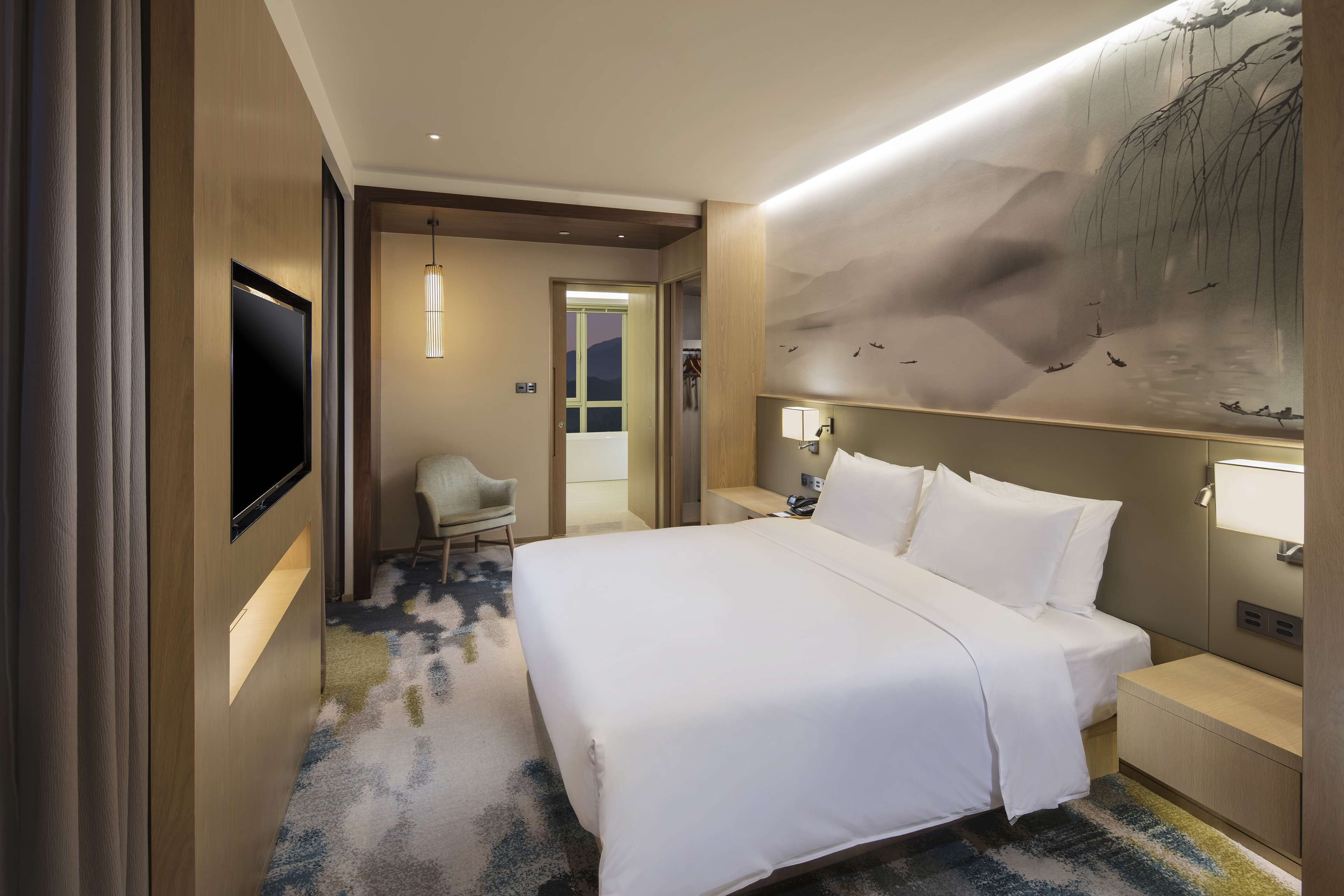 A hotel room with a large bed, a desk, a chair, a TV, and a wall with a nature mural.