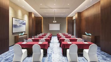 Un salón de conferencias con filas de mesas y sillas, una pantalla de proyector y un cuadro grande en la pared.