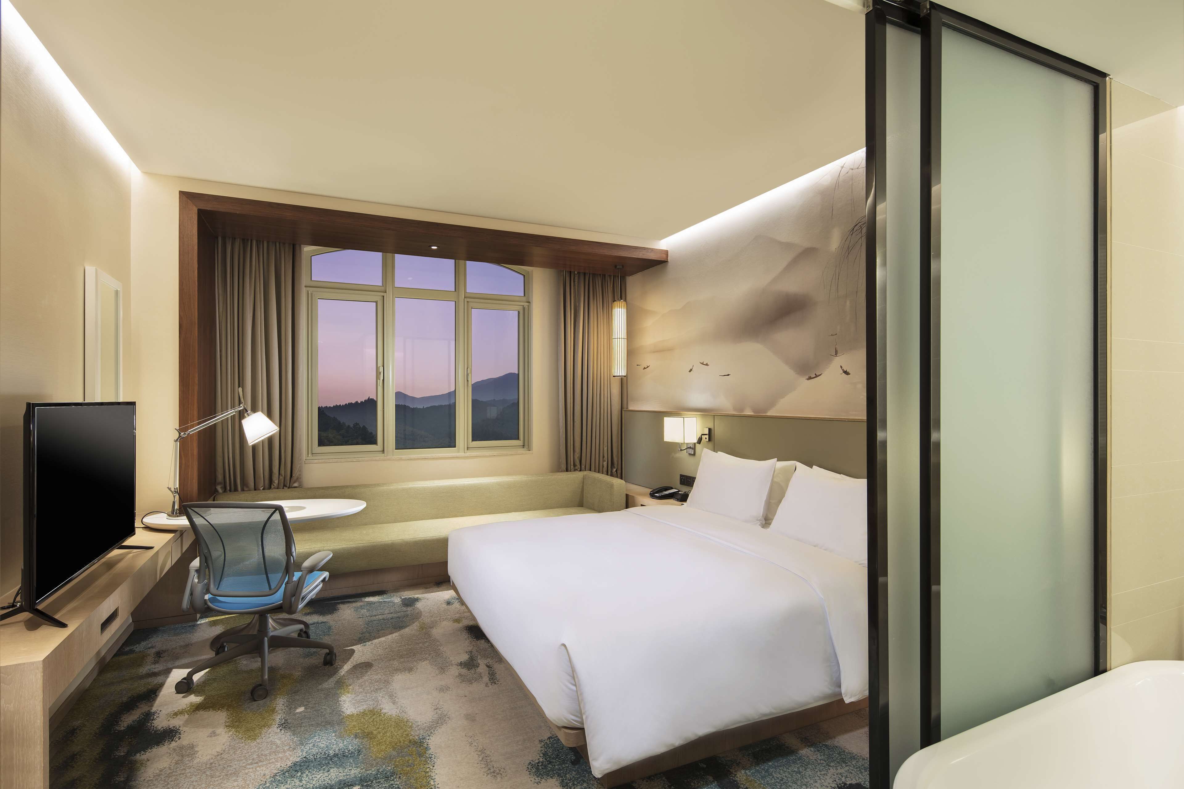 A hotel room with a bed, a desk with a computer, a chair, a TV, a window with curtains, and a view of the mountains.