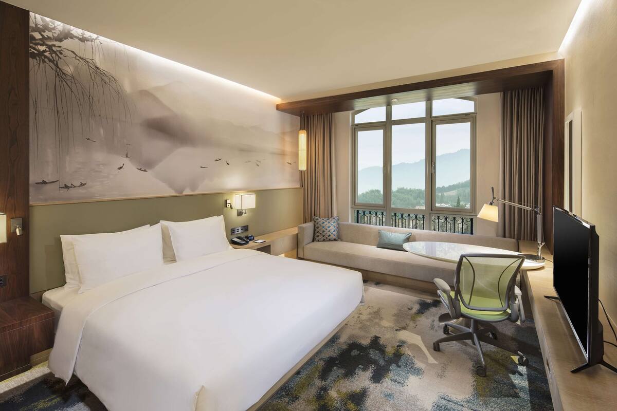 Ein Hotelzimmer mit einem großen Bett, einer Couch, einem Schreibtisch mit Stuhl, einem Fernseher und Bergblick durch ein großes Fenster.