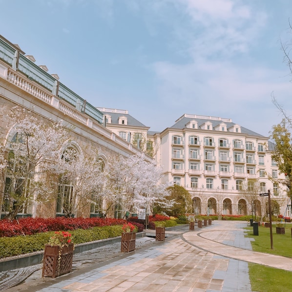 A large, classical building with symmetrical architecture, a central courtyard with a fountain, and a well-maintained garden with flowering trees and red foliage.