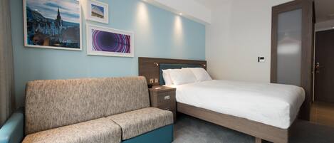 A hotel room with a bed, a sofa, a nightstand, and wall art.