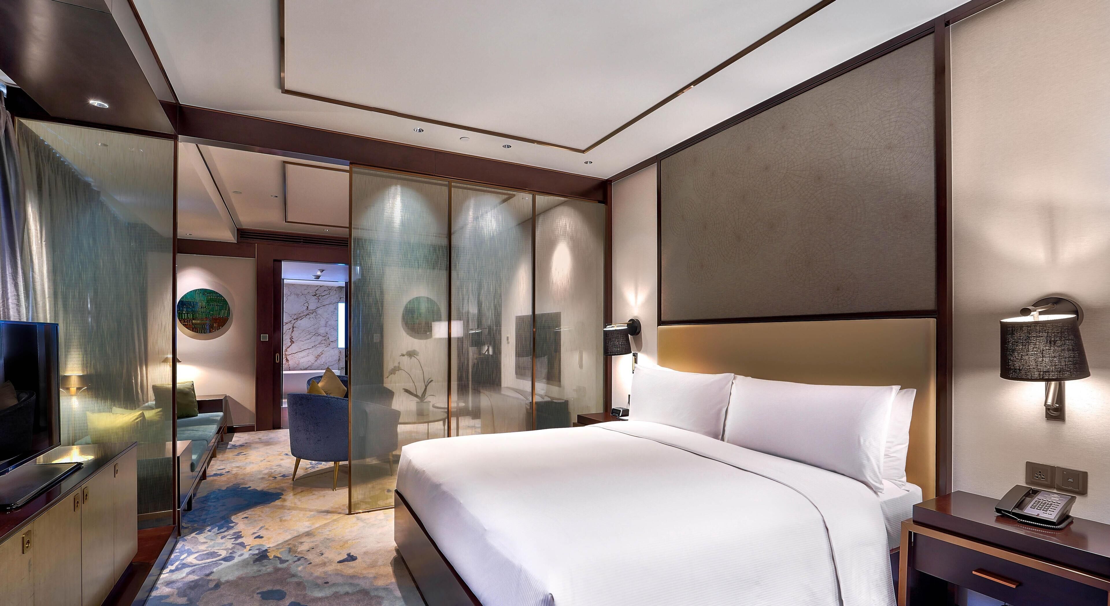 Habitación de hotel con una cama grande, un escritorio con computadora, un televisor y un divisor de vidrio.